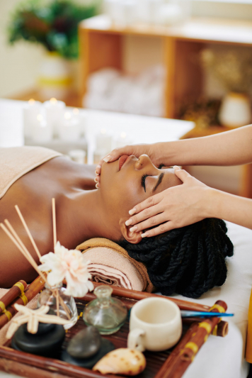 Beautiful young Black woman enjoying face and head massage in spa salon when lying on bed next to tray with aroma oils